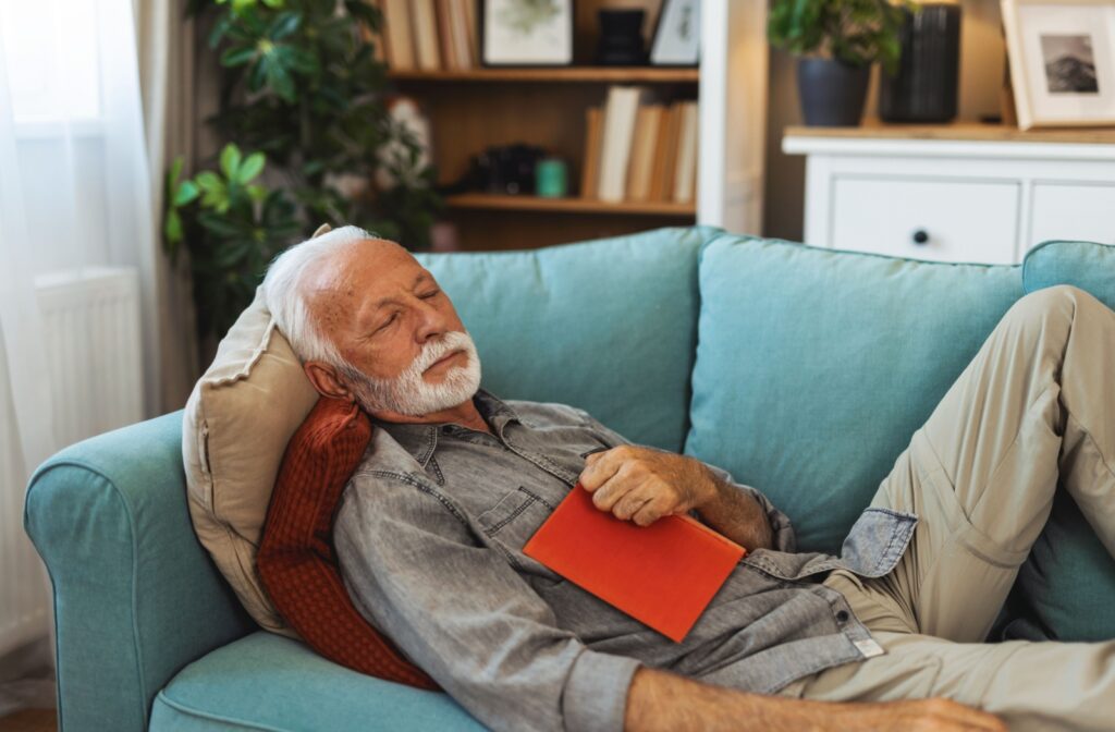 An older adult sleeping peacefully on the couch in the middle of the day, while holding a book.