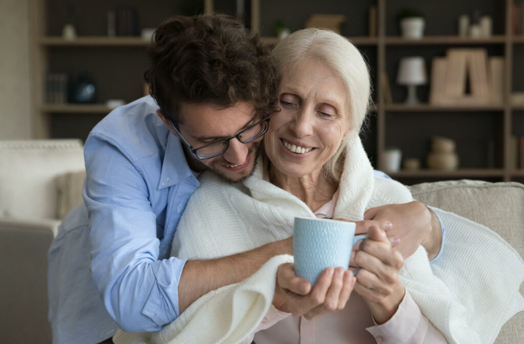 An adult child hugging their older parent in assisted living after bringing them a mug of coffee.
