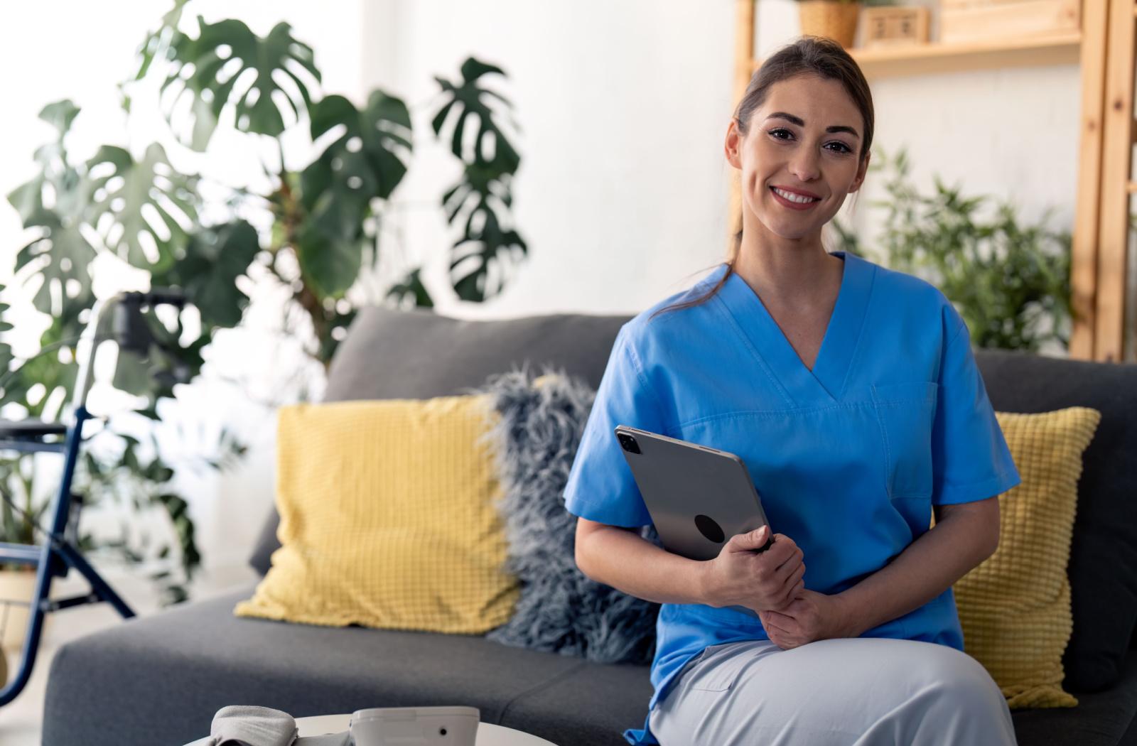 A respite caregiver smiles while holding a tablet computer and sitting on a gray sofa.
